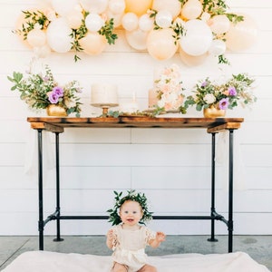 Może przedstawiać: Mała dziewczynka w białej sukience i zielonej koronie z liści siedzi na białym dywanie przed drewnianym stołem ozdobionym kwiatami, balonami i ciastami.