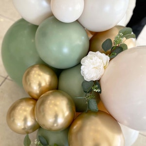 Puede incluir: Una guirnalda de globos verdes, dorados y crema con una flor blanca y detalles de vegetaci&oacute;n.