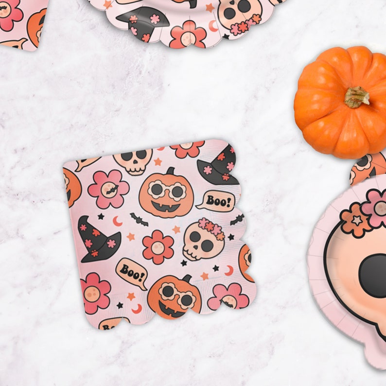 May include: Pink paper napkins with a Halloween theme. The napkins feature a repeating pattern of pumpkins, skulls, witches hats, and flowers. The text "Boo!" appears on the napkins.