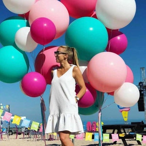 Op de afbeelding: Een vrouw in een witte jurk staat voor een grote groep kleurrijke ballonnen. De ballonnen zijn voornamelijk roze, wit en turquoise.