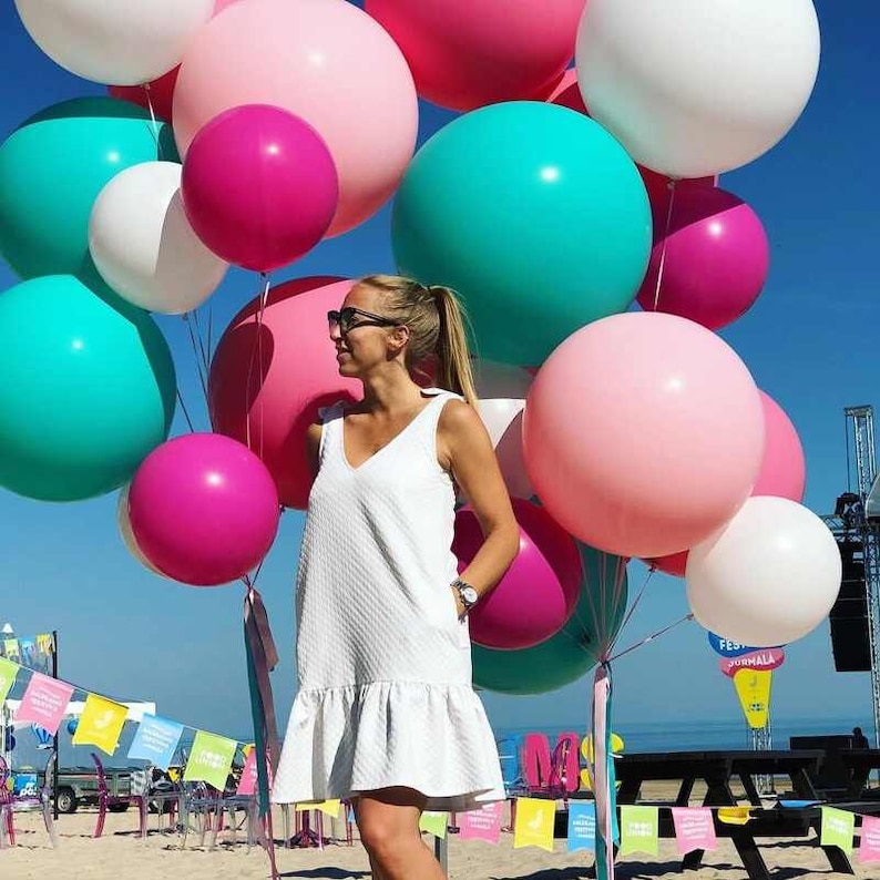 Op de afbeelding: Een vrouw in een witte jurk staat voor een grote groep kleurrijke ballonnen. De ballonnen zijn voornamelijk roze, wit en turquoise.