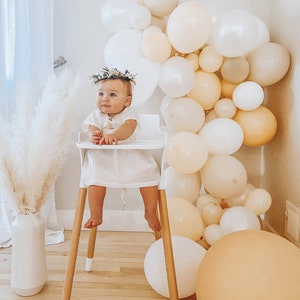 May include: A baby girl wearing a white dress and a floral crown sits in a white high chair. The chair has wooden legs and a white tray. The baby is surrounded by a garland of cream and beige balloons.