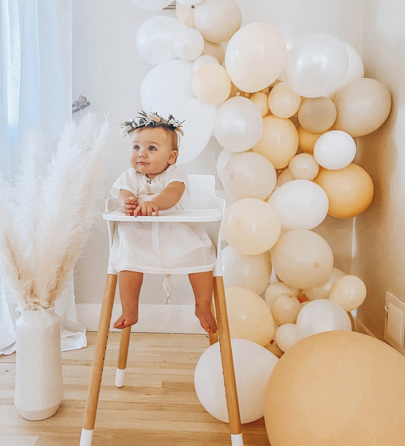 May include: A baby girl wearing a white dress and a floral crown sits in a white high chair. The chair has wooden legs and a white tray. The baby is surrounded by a garland of cream and beige balloons.