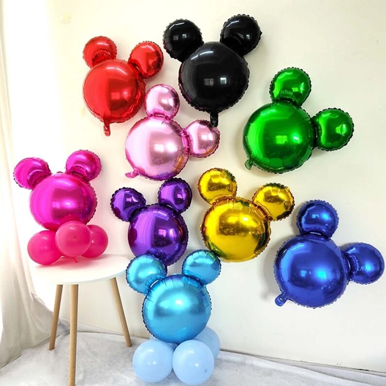 May include: Assortment of eight Mickey Mouse shaped foil balloons in various colors, including red, black, pink, green, magenta, purple, gold, and blue. The balloons are arranged on a white wall and a table.