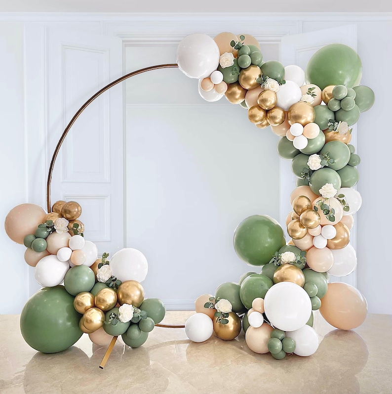 May include: A gold metal hoop arch decorated with green, white, and gold balloons. The balloons are arranged in a garland shape and are tied together with white ribbon. The arch is standing on a white surface.