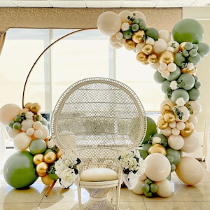 Puede incluir: Una silla de pavo real de mimbre blanco con un arco de metal dorado decorado con globos verdes, blancos y dorados.