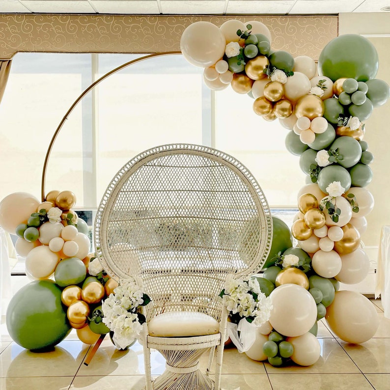Puede incluir: Una silla de pavo real de mimbre blanco con un arco de metal dorado decorado con globos verdes, blancos y dorados.
