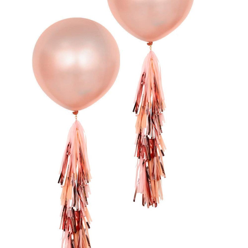 May include: Two rose gold balloons with matching tassel tails. The balloons are round and shiny, with a metallic finish. The tassels are made of strips of pink, rose gold, and copper-colored paper. The balloons are tied with a small heart-shaped detail.