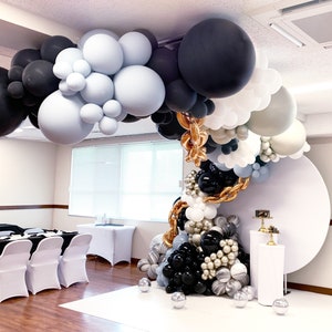 Puede incluir: Un arco de globos hecho de globos negros, blancos y azul claro. El arco est&aacute; decorado con cinta dorada y est&aacute; instalado frente a un fondo blanco.
