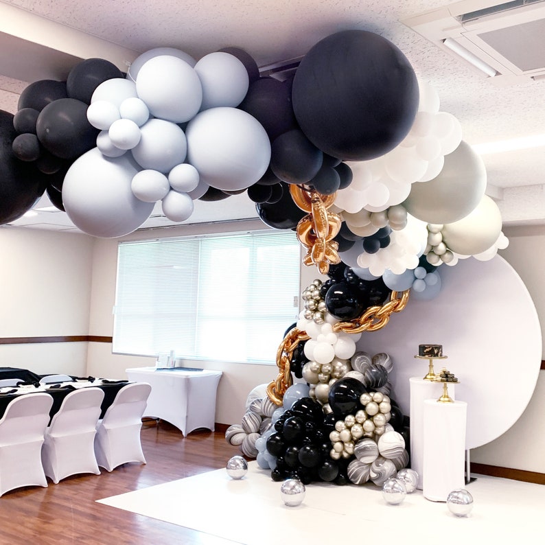 Puede incluir: Un arco de globos hecho de globos negros, blancos y azul claro. El arco est&aacute; decorado con cinta dorada y est&aacute; instalado frente a un fondo blanco.