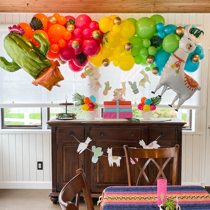 May include: A llama balloon with a colorful balloon garland, a cactus balloon, and a table setting with a llama themed banner and a colorful tablecloth.