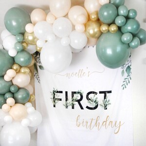 May include: A white banner with green leaves and gold lettering that says "noelle's first birthday". A garland of green, white, and gold balloons is draped over the top of the banner.