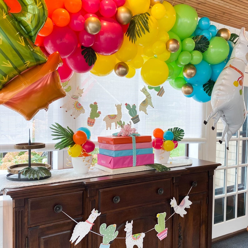 May include: A colorful balloon garland with rainbow, yellow, green, and blue balloons. The garland is decorated with green leaves and paper llama cutouts. A wooden table with a stack of wrapped gifts sits in front of the garland.