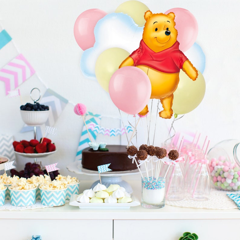 May include: A Winnie the Pooh balloon centrepiece with pink, yellow and white balloons. The balloons are arranged around a Winnie the Pooh balloon, which is holding a red balloon. The balloons are on a table with a cake, cupcakes and other decorations.