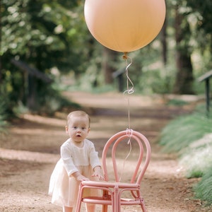 May include: A young child in a white dress stands next to a pink chair with a large peach-colored balloon floating above.