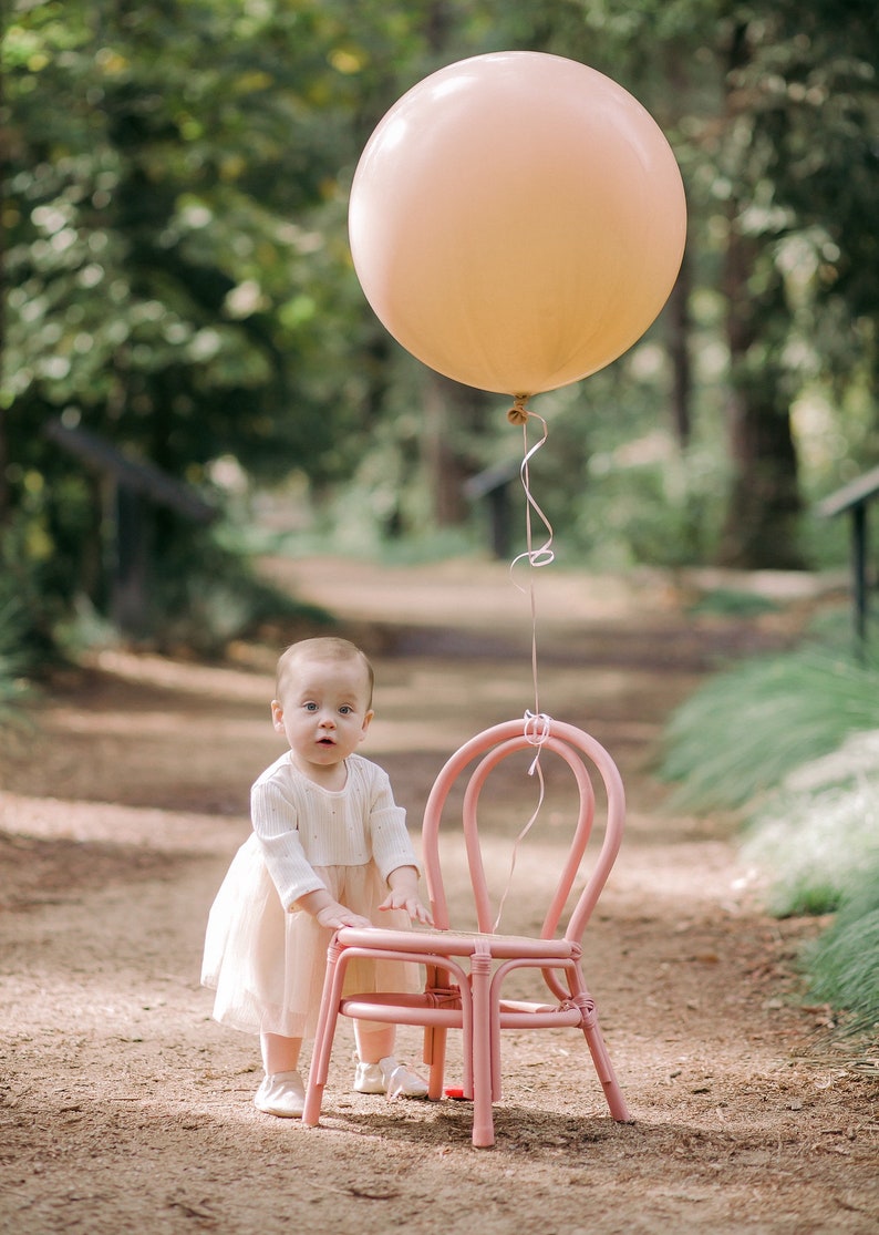 May include: A young child in a white dress stands next to a pink chair with a large peach-colored balloon floating above.
