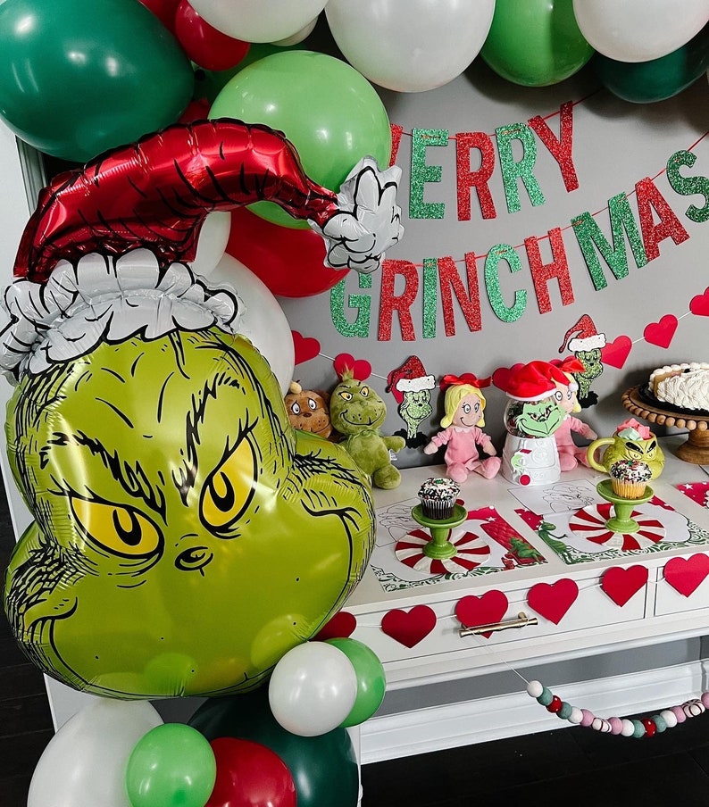 Pu&ograve; includere: Un palloncino verde Grinch con un cappello rosso e un bordo di pelliccia bianca. Il palloncino &egrave; circondato da palloncini verdi, rossi e bianchi. Uno striscione di glitter verde e rosso recita "Merry Grinchmas".