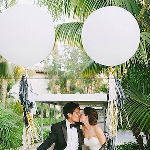 May include: A couple in a white golf cart decorated with black and gold streamers, kissing under two large white balloons.