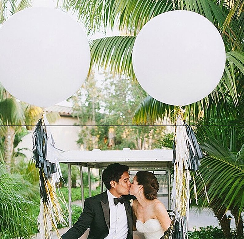 May include: A couple in a white golf cart decorated with black and gold streamers, kissing under two large white balloons.