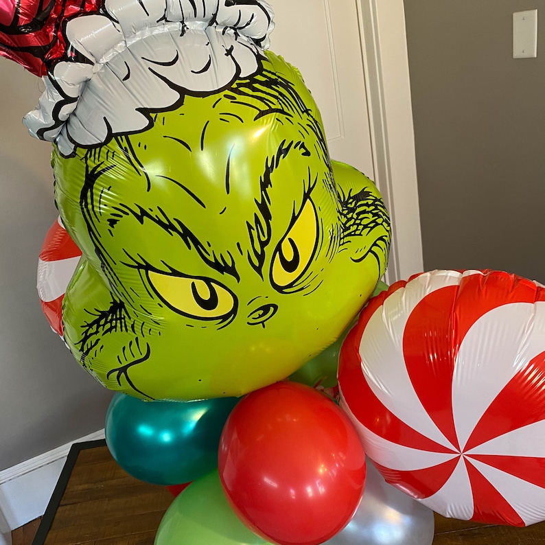 Pu&ograve; includere: Un palloncino verde Grinch con un cappello di Babbo Natale bianco, circondato da palloncini a forma di bastoncino di zucchero rosso e bianco e un palloncino verde, rosso e argento.