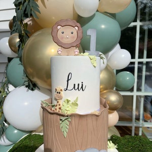 May include: A two-tiered cake decorated with a brown lion, a giraffe, and green leaves. The top tier is white with the name "Lui" written in black. The bottom tier is brown and resembles a tree trunk. The cake is surrounded by balloons in various colors.
