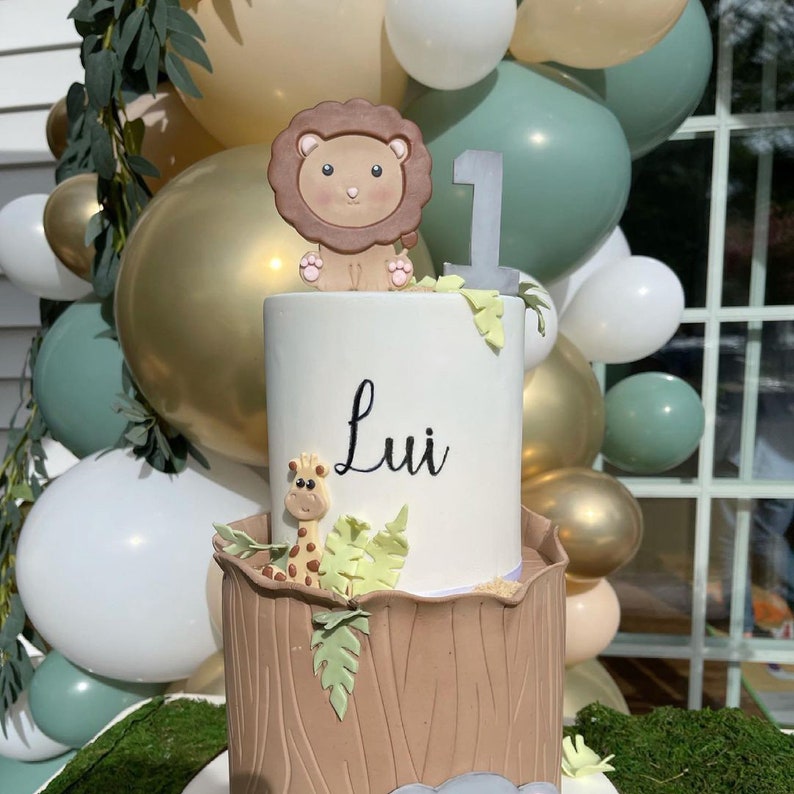 May include: A two-tiered cake decorated with a brown lion, a giraffe, and green leaves. The top tier is white with the name "Lui" written in black. The bottom tier is brown and resembles a tree trunk. The cake is surrounded by balloons in various colors.