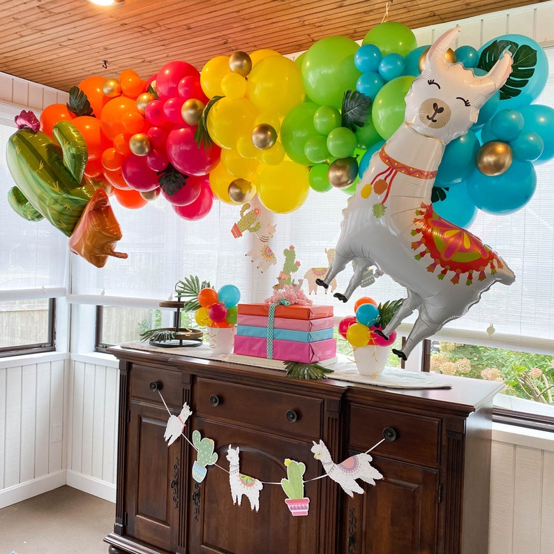 May include: A white llama balloon with colorful accents, surrounded by a garland of colorful balloons. The balloons are in shades of red, orange, yellow, green, and blue. The llama balloon is holding a banner with the text "Happy Birthday".