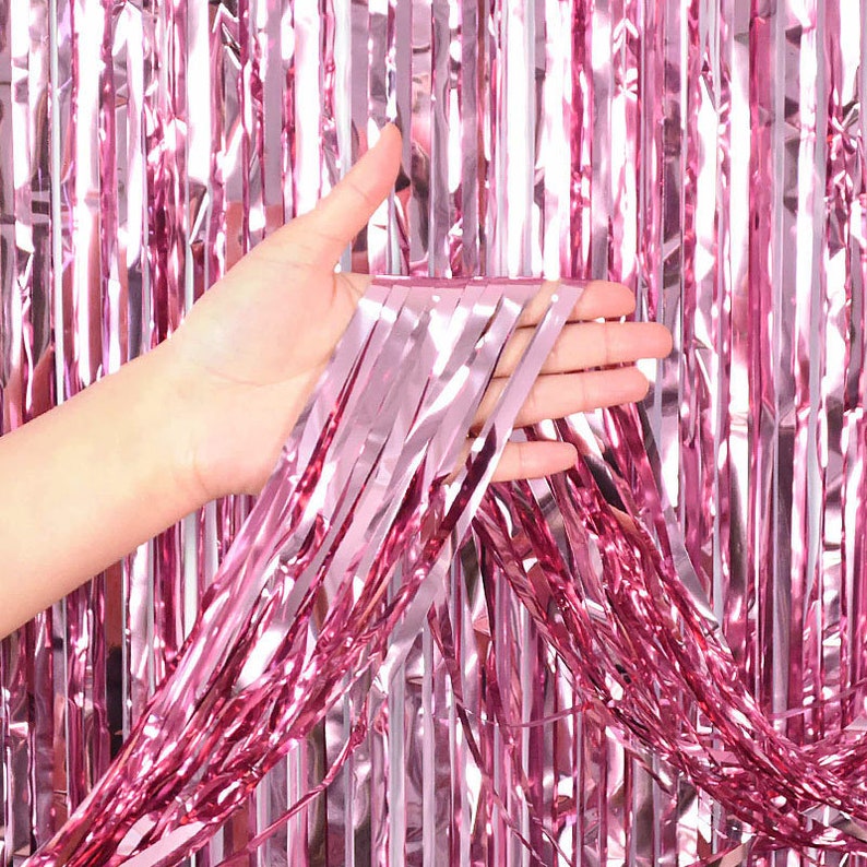 May include: Pink metallic fringe curtain. A hand holds a section of the curtain, showcasing its shiny, reflective surface. The curtain is made of vertical strips, creating a shimmering effect, suitable for party decorations or photo backdrops.
