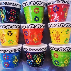 May include: A collection of nine small ceramic flower pots arranged in a three-by-three grid. Each pot features a white rim with a black swirling pattern and a vibrant, multi-colored body decorated with raised, hand-painted floral motifs.