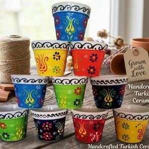 May include: A collection of ten small ceramic pots arranged in a pyramid, featuring vibrant colors including blue, yellow, red, green, and black. Each pot is decorated with hand-painted floral patterns and a black swirling border at the rim.