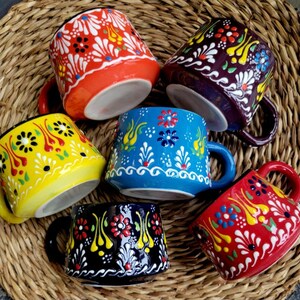 May include: Six colorful ceramic mugs with floral patterns. The mugs are in various colors, including red, yellow, blue, purple, and black, each with a unique design. The mugs are arranged on a woven, textured surface.