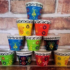 May include: A collection of nine ceramic flower pots stacked in a pyramid formation. Each pot features a vibrant solid color base, including blue, yellow, red, green, and black, decorated with hand-painted floral motifs and a black swirling rim.