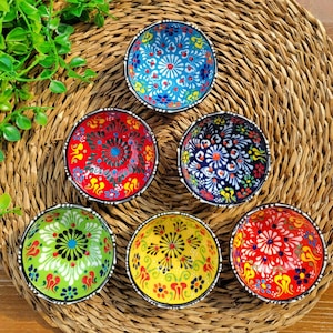 May include: Six small ceramic bowls with colourful floral patterns. The bowls are arranged in a circle on a woven straw mat. The bowls are all different colours, including blue, red, green, and yellow.