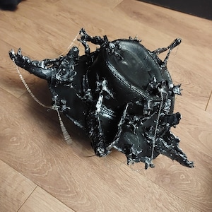 Könnte beinhalten: Ein schwarzes, skulpturales Kopfstück mit einem komplizierten, strukturierten Design. Das Kopfstück weist eine komplexe Anordnung von stacheligen Vorsprüngen auf und ist mit silberfarbenen Kettendetails versehen. Das Stück liegt auf einem Holzboden.