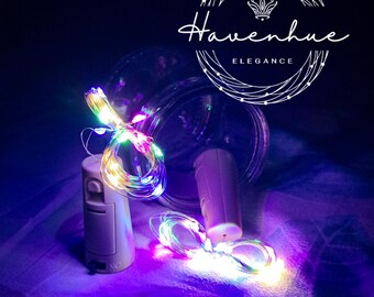 May include: Two white battery-operated bottle stoppers with multi-coloured fairy lights wrapped around them. The lights are on and glowing. The stoppers are on a purple fabric surface. The text "Havenhue Elegance" is visible in the background.