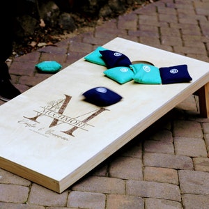 Könnte beinhalten: Ein personalisiertes Holz-Cornhole-Spiel mit dem Nachnamen "Muchmore" und den Namen "Layla & Brennan", die auf das Brett graviert sind. Das Brett ist auf einer gepflasterten Terrasse mit blauen und türkisfarbenen Wurfsäcken aufgestellt.