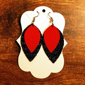 Pode incluir: Um par de brincos pendentes com um design em camadas vermelho e preto. Os brincos têm a forma de uma lágrima e têm um gancho prateado.