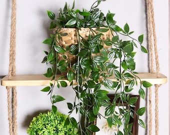 May include: Two wooden shelves hanging from a rope, with a potted plant on each shelf. The top shelf has a green plant in a woven basket, and the bottom shelf has a green plant in a white pot. A framed picture is on the top shelf.