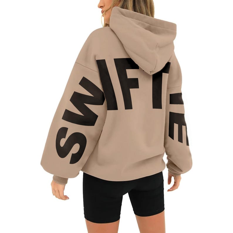May include: A tan hoodie with a large black "SWIFTIE" graphic printed on the back. The hoodie has a drawstring hood and long sleeves.