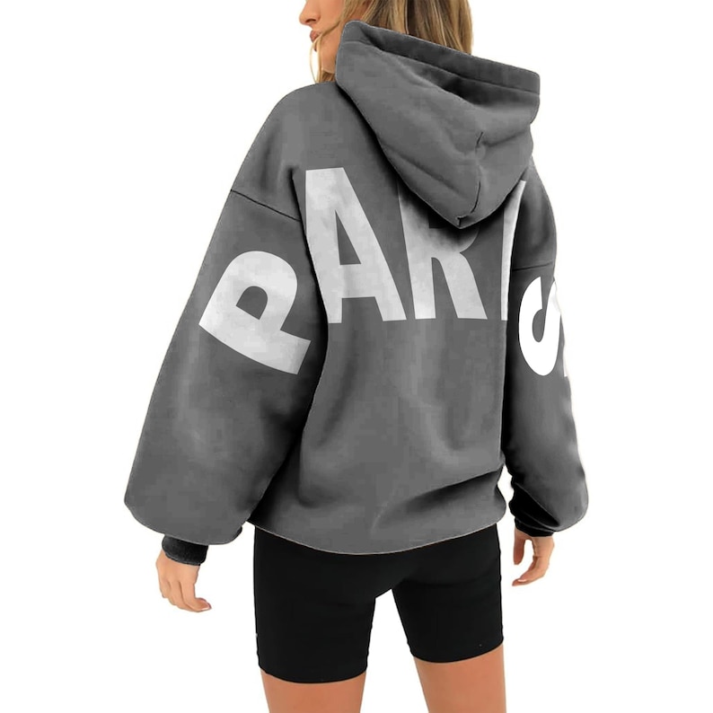 May include: A gray hooded sweatshirt with the word "PARIS" printed in white on the back. The sweatshirt has a relaxed fit and a drawstring hood.