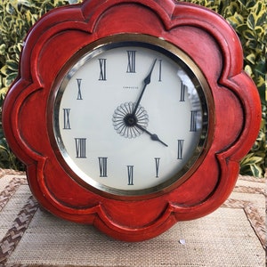 Puede incluir: Un reloj de madera rojo con un marco en forma de flor y n&uacute;meros romanos en la esfera. La esfera del reloj es blanca con detalles dorados y el nombre de la marca "Caravelle" est&aacute; impreso en la esfera.