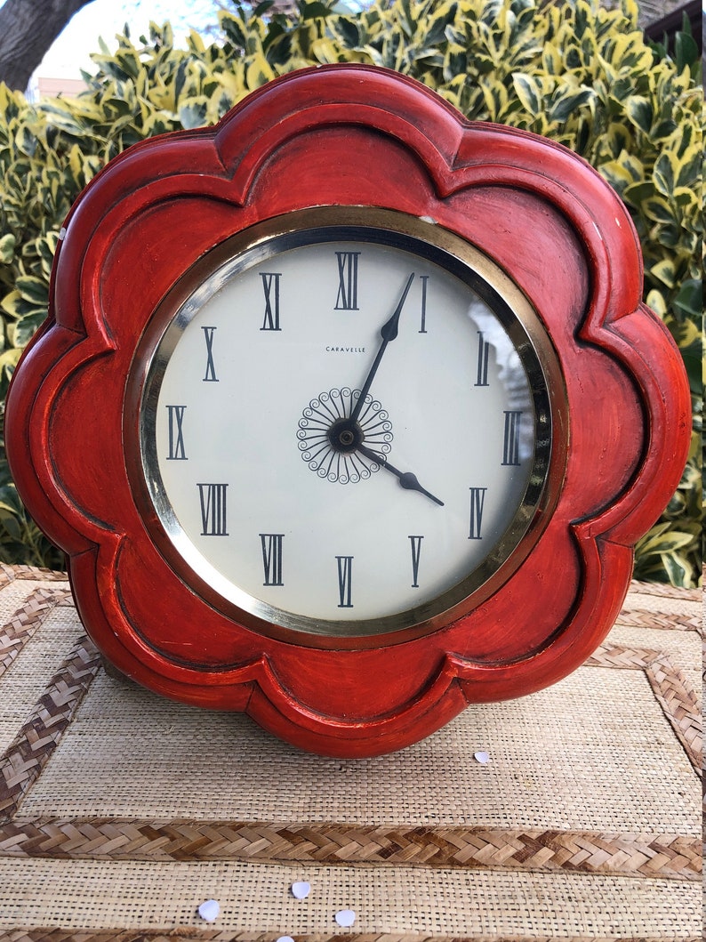 Puede incluir: Un reloj de madera rojo con un marco en forma de flor y n&uacute;meros romanos en la esfera. La esfera del reloj es blanca con detalles dorados y el nombre de la marca "Caravelle" est&aacute; impreso en la esfera.