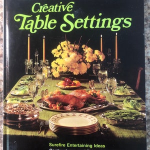 May include: A book titled "Creative Table Settings" by Better Homes and Gardens. The cover features a beautifully set table with a centerpiece of yellow flowers, candles, and a roasted chicken. The book offers ideas for entertaining, centerpieces, table decorations, and how to select tableware.