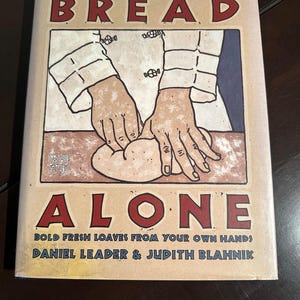 May include: A cookbook titled "BREAD ALONE" with a vintage-style illustration of hands kneading dough. The cover features the title in red block letters, the author's names, and the tagline "BOLD FRESH LOAVES FROM YOUR OWN HANDS".