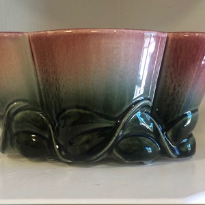 May include: A pink and green ceramic planter with a flower design. The planter has a scalloped rim and a wide base.