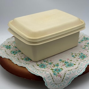 May include: A rectangular, cream-colored container with a matching lid. The container is sitting on a floral patterned handkerchief with a scalloped edge. The handkerchief is on a round, wooden tray.
