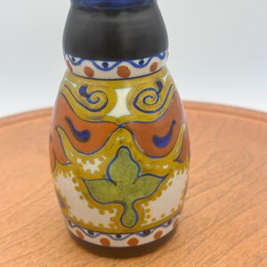 May include: A small ceramic vase with a dark brown neck and base. The body features a cream-colored background decorated with hand-painted patterns in mustard yellow, burnt orange, olive green, and cobalt blue, including leaf and scroll motifs.