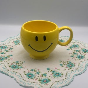 May include: A yellow ceramic mug with a black smiley face design. The mug is sitting on a white and blue floral patterned fabric.