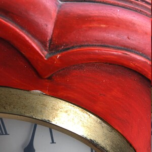 Puede incluir: Un reloj rojo con un borde dorado y una esfera blanca. La esfera del reloj tiene n&uacute;meros romanos y la palabra "Aravelle" impresa en ella.