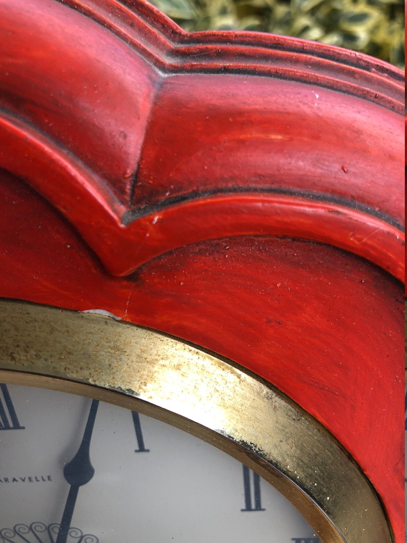 Puede incluir: Un reloj rojo con un borde dorado y una esfera blanca. La esfera del reloj tiene n&uacute;meros romanos y la palabra "Aravelle" impresa en ella.